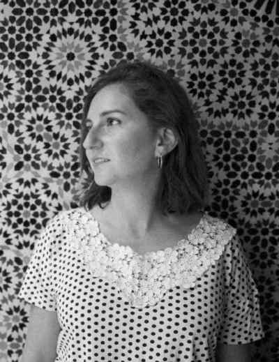 La imagen es un retrato en blanco y negro de una mujer con el pelo castaño medio, mirando hacia la izquierda. Lleva un vestido de lunares blancos y negros con un cuello de encaje blanco. El fondo es una pared con un patrón repetitivo de mosaicos florales en tonos oscuros. La expresión de la mujer es seria y contemplativa.