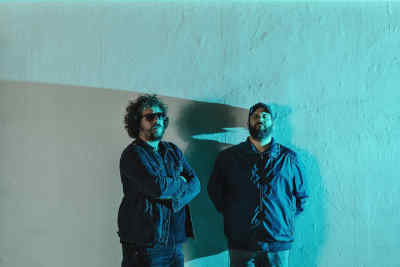 Dos hombres con aspecto informal posan frente a una pared de color turquesa claro, con iluminación suave y contrastada.