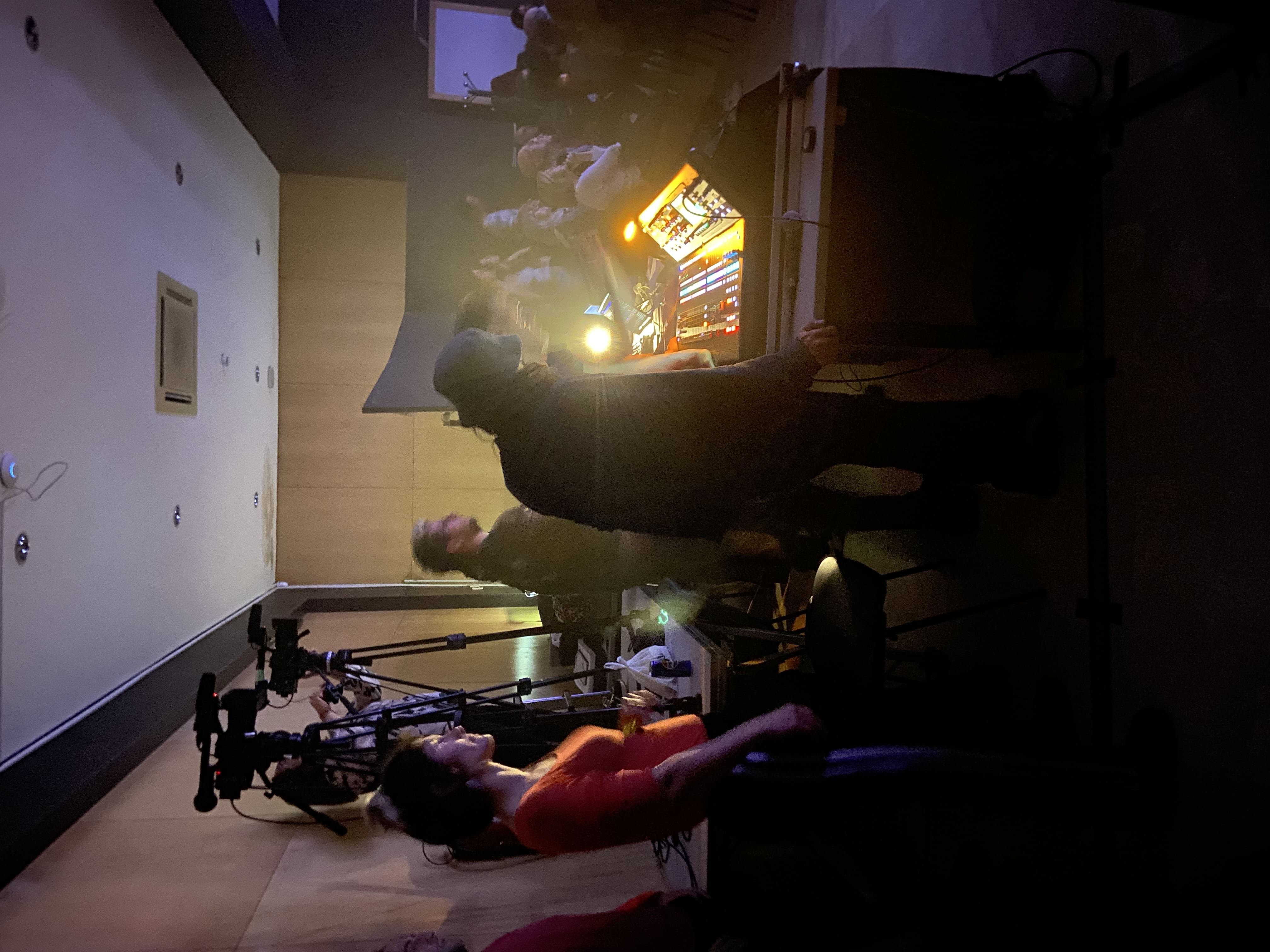 En el centro de la fotografía (hehca en vertical) se recorta la silueta de una persona con un sombreo que está dirigiendo su cuepor y su mano hacia la mesa de sonido que ocupa la parte central drecha de la fotograíd. esta rodeado de cámaras, más técnicos y al fondo a la derecha se ve el público que está asistiendo a unos de los conciertos que tiene lugar en la sala Plató del Rey Chico