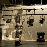 Técnico de iluminación teatral trabajando delante de una pantalla translúcida. Está rodeado de focos y parece estar revisando o ajustando algo relacionado con la iluminación del escenario.