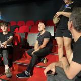 El profesor sentado en la escaleras de patio de butacas del teatro rodeado de un grupo de alumnos. Todos vestidos de negro con el suelo, escaleras y butacas en rojo