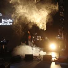 La imagen muestra un escenario con una atmósfera de luces y humo. Hay micrófonos y soportes de partituras; uno de ellos tiene una hoja colocada. Se ve una persona en el escenario, y el fondo muestra el logo de 