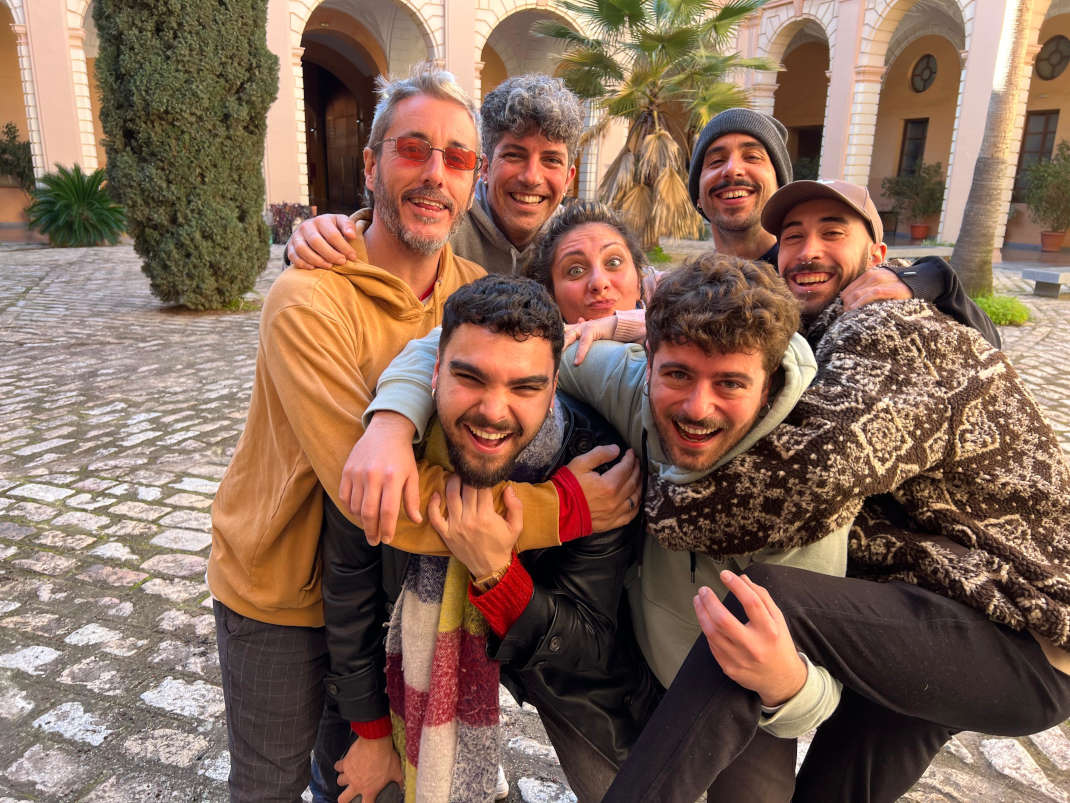 La imagen muestra a un grupo de ocho jóvenes, aparentemente amigos, abrazándose y riéndose en el patio de lo que parece ser un edificio antiguo con arcos. Muchos están llevando a otros sobre sus hombros en una demostración de jovialidad y camaradería. La atmósfera es alegre y despreocupada.