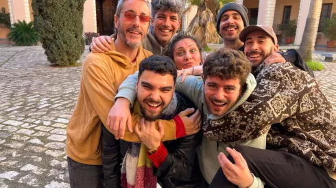 La imagen muestra a un grupo de ocho jóvenes, aparentemente amigos, abrazándose y riéndose en el patio de lo que parece ser un edificio antiguo con arcos. Muchos están llevando a otros sobre sus hombros en una demostración de jovialidad y camaradería. La atmósfera es alegre y despreocupada.