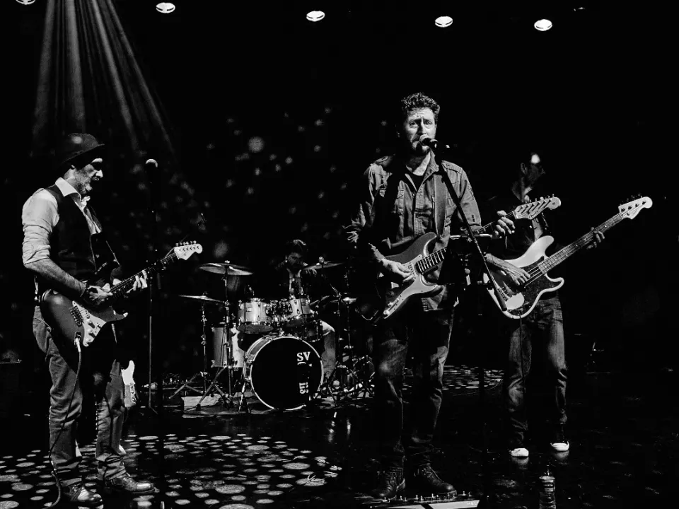 Fotografía en blanco y negro de una banda tocando en un escenario. Se ven tres músicos claramente: un guitarrista principal con sombrero, un vocalista/guitarrista que está al frente y un bajista. Hay un baterista parcialmente visible detrás de ellos. La iluminación del escenario, con luces que apuntan hacia abajo, crea un ambiente dramático. La escena evoca una presentación musical en vivo en un ambiente oscuro y de club.
