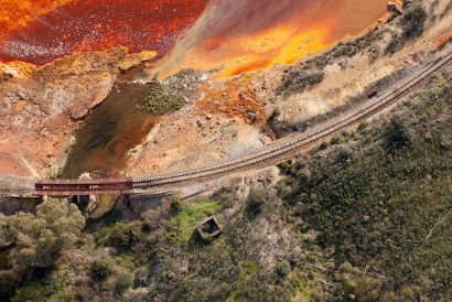 Vista aerea de Río Tinto: paisaje dividido por la via del tren. A un lado zona de vegetación y al otro una masa de agua de color anaranjado.