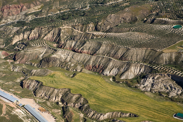 Fotografía aerea de montañas. Se pueden apreciar cultivos de olivo y unas naves.