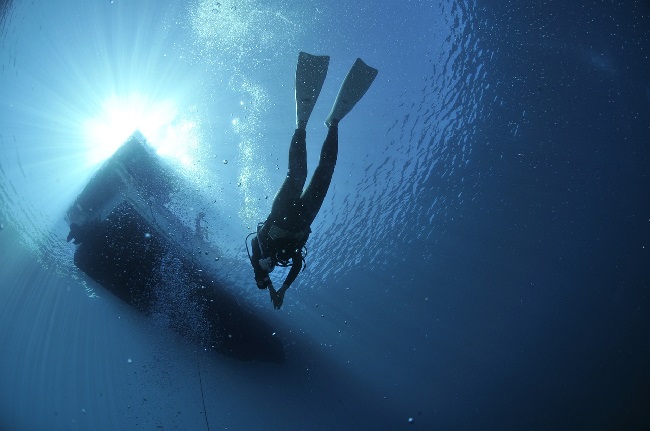 Buzo sumergido en el mar. Sobre él se transparenta la silueta de una barca, en la superficie