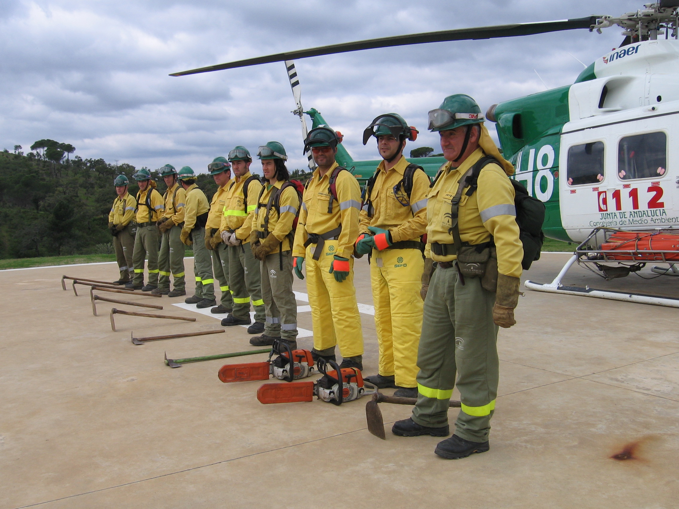 Ampliar imagen: operarios del Plan INFOCA con uniforme y equipo de trabajo junto a un helicóptero de transporte y extinción de incendios