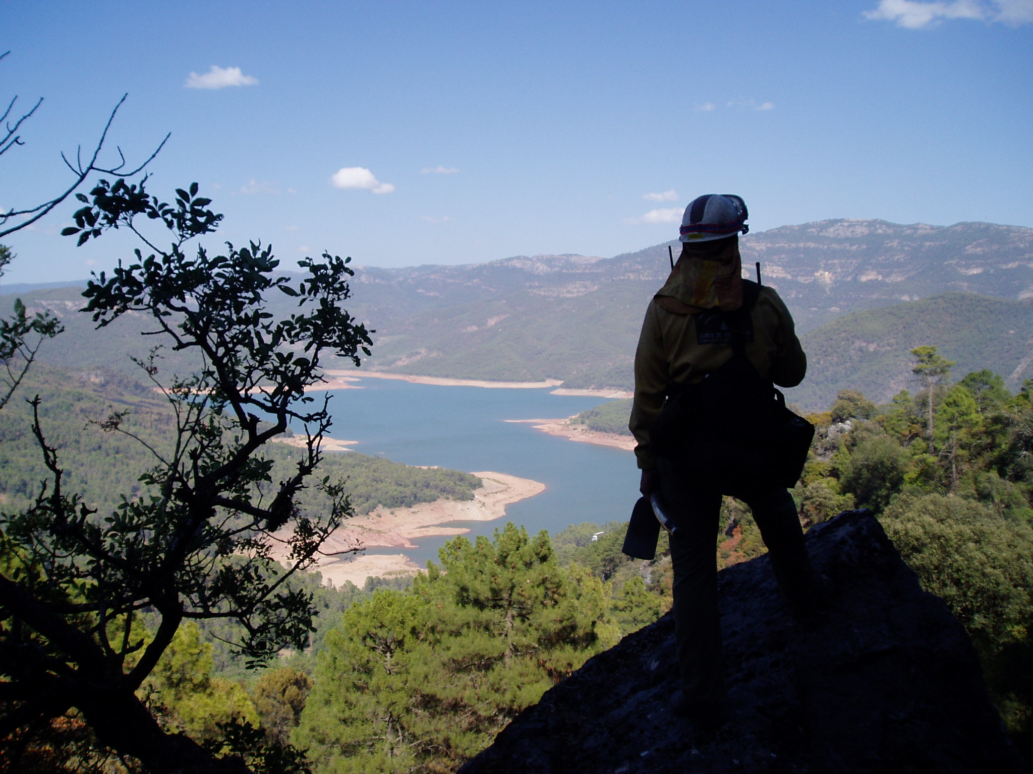 Ampliar imagen: silueta de un bombero forestal avistando desde lo alto el estado de las masas boscosas y vegetación que rodean a un embalse.