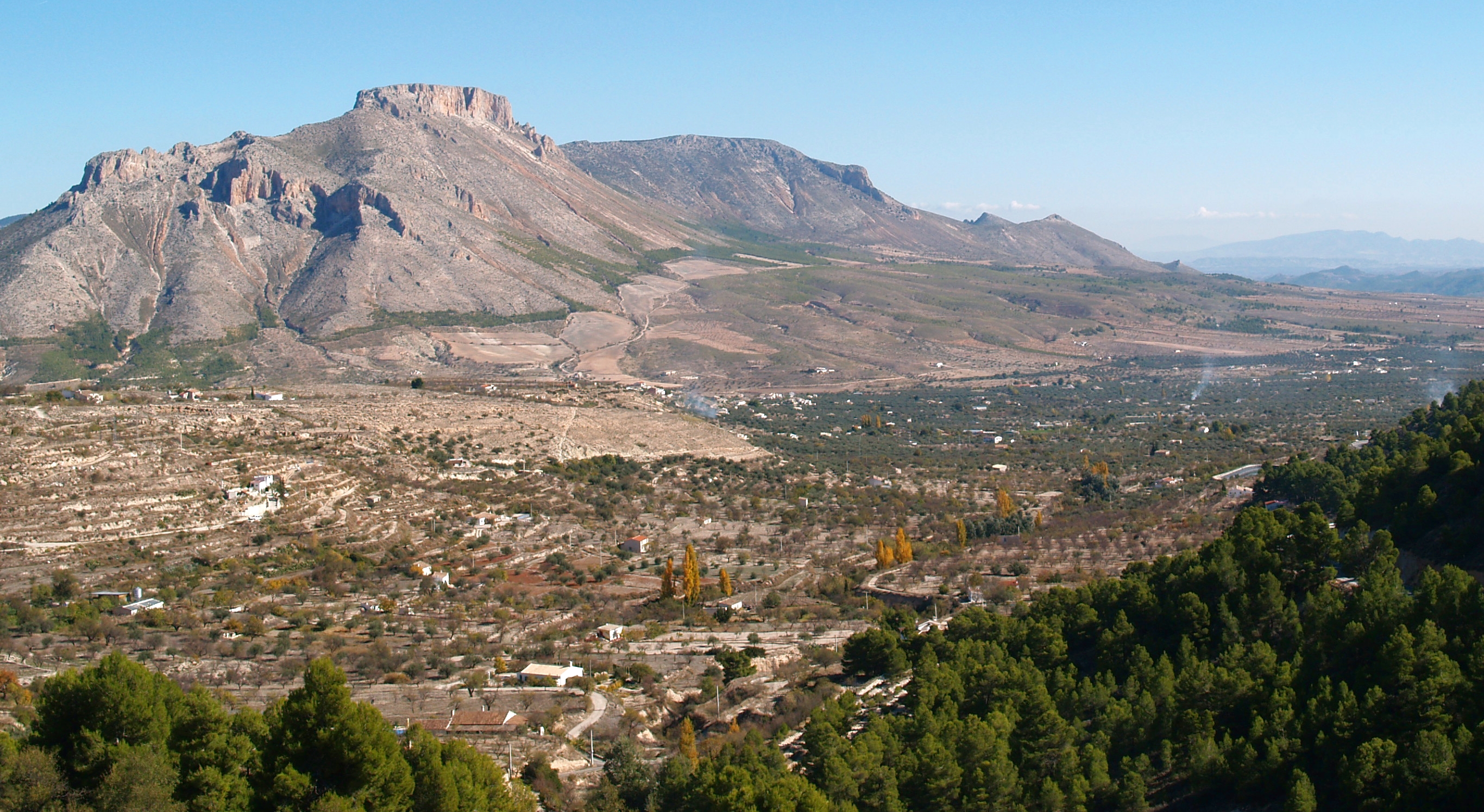 Ampliar imagen: cumbre rocosa que a través de una ladera va perdiendo pendiente pasando a un paisaje llano en el que se observan cultivos y la presencia arbórea de pinar.