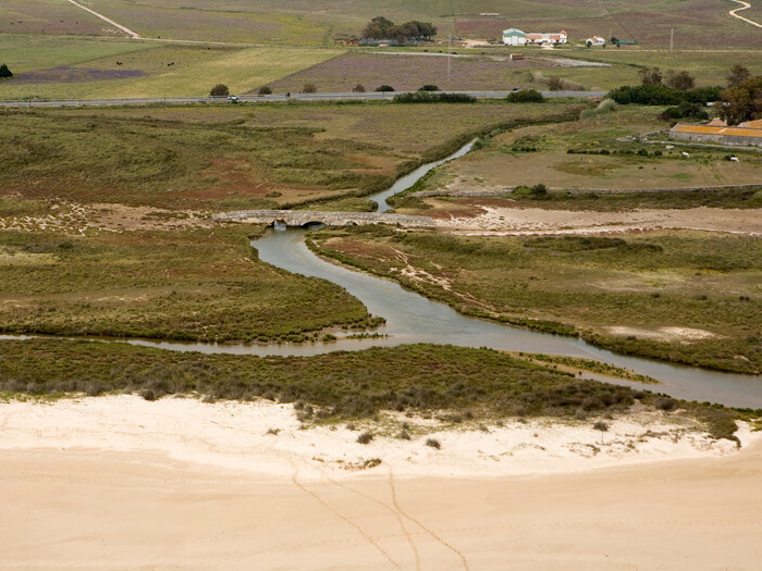 Río serpenteante con un puente de piedra rústico, desenboca en una playa de arena clara y campos verdes