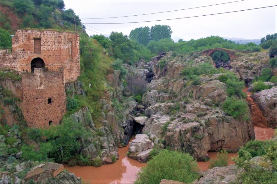 Ampliar imagen: Ampliar imagen: ruinas de piedra junto a un río rojizo en un cañón rocoso, con vegetación y un puente antiguo