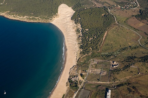 Fotografía aérea de una playa formada por dunas de arena fina, mar de color azul intenso un yacimiento arqueológico y vegetación