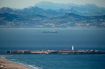 Vista aérea de una isla rocosa con un faro, un barco en el agua y montañas al fondo