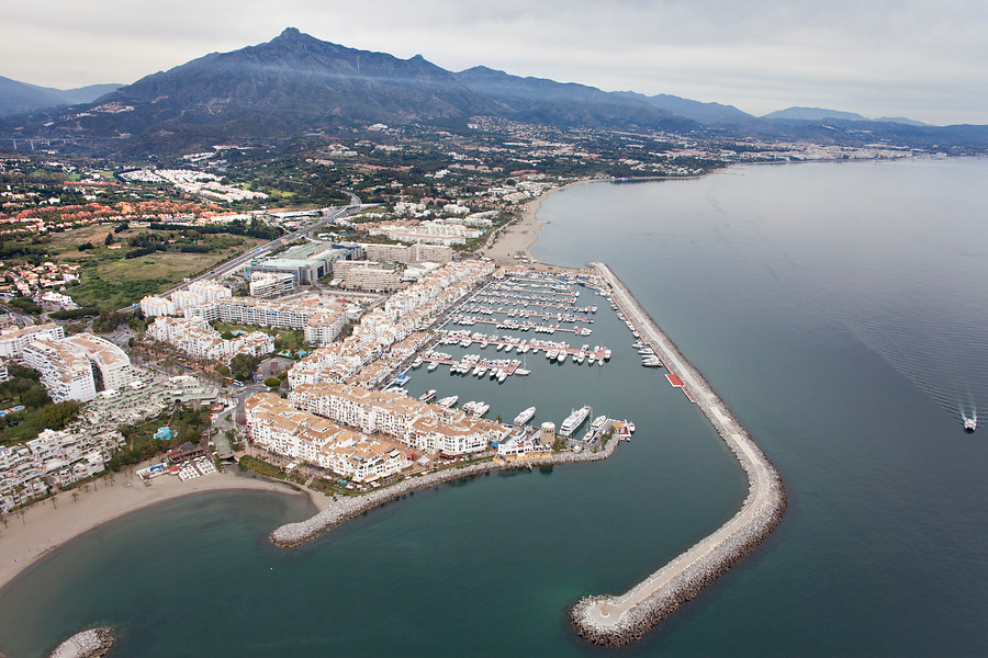 Ampliar imagen: Vista de pájaro de una zona urbana con puerto deportivo y paisaje de montaña al fondo.