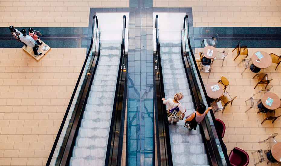 Clientes de un centro comercial bajando por las escaleras mecánicas a la planta de moda y restauración.
