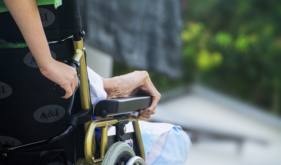 Una trabajadora de una residencia ayuda a una anciana en silla de ruedas a desplazarse por el centro.