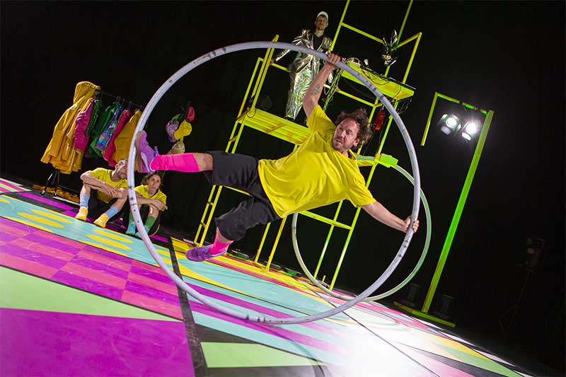 Artista haciendo una acrobacia dentro de un aro en un escenario colorido con luces y otros performers al fondo